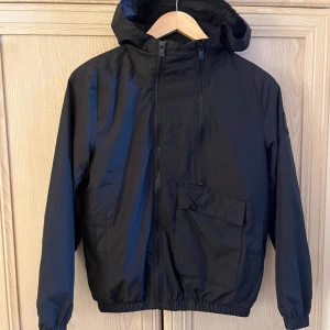 Svart vindjacka med huva Lyle & Scott 10-11 år storlek  - Storlek 10-11 år.Säljer en svart vindjacka från Lyle & Scott. Den har en praktisk huva och dragkedja framtill. Jackan är perfekt för blåsiga dagar och har en cool logga på ärmen. Den är i mycket bra skick och passar perfekt för vår och höst.