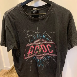 Svart AC/DC T-shirt - Cool svart AC/DC T-shirt med tryck från High Voltage Tour 1976. Den har ett elektriskt mönster med bandets logga i rött och blått på framsidan och turnédatum på baksidan. Perfekt för rockälskare! 🖤