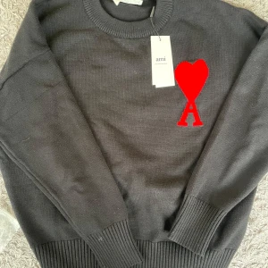 Svart stickad tröja från Amish - Säljer en snygg svart stickad tröja från Ami med ett stort rött hjärta på bröstet. Tröjan har en klassisk rund hals och långa ärmar med ribbade muddar. Perfekt för både höst och vinter, och den är i mycket bra skick. Passar perfekt för en stilren look!