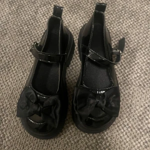 Svarta Mary janes skor med en rosett på - Säljer ett par svarta Mary janes skor i syntetmaterial med en söt rosett framtill. De har en robust sula och justerbart spänne för perfekt passform. Perfekta för både vardag och festliga tillfällen!
