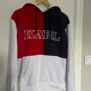 Flerfärgad hoodie från Karl Kani - Säljer en cool flerfärgad hoodie från Karl Kani. Den har en unik design med röd, svart och vit färgblock. Stora bokstäver 'KARL' och 'KANI' pryder framsidan och baksidan. Perfekt för en avslappnad stil med en stor ficka framtill och justerbar huva. Har någon mindre defekt, se bilder.