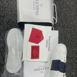 Valentino Garavani sneakers - Säljer ett par snygga Valentino Garavani sneakers i vitt med en bred mörkblå rem över. Skorna är i nytt skick och kommer med originalförpackning och påse. Perfekta för en stilren look!