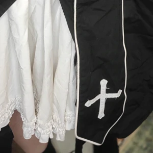 Vit och svart kjol med korsdetaljer lolita inspirerad  - Säljer en unik vit och svart kjol med coola korsdetaljer. Kjolen har en volangkant och är perfekt för en edgy look. Den är i mycket bra skick och passar perfekt för höst och vinter. Perfekt för dig som vill sticka ut! En lolita inspirerad outfit 