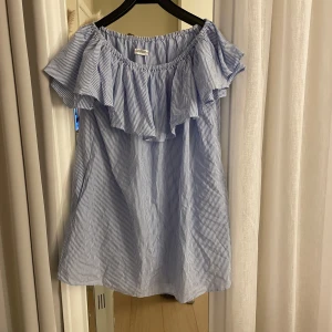 Blå- och vitrandig offshoulder klänning - Söt blå- och vitrandig offshoulder klänning med volangdetaljer. Perfekt för sommardagar eller en avslappnad fest. Klänningen har en luftig passform och är tillverkad i ett lätt och bekvämt material. Osäker på storlek men passar mej som har xs s