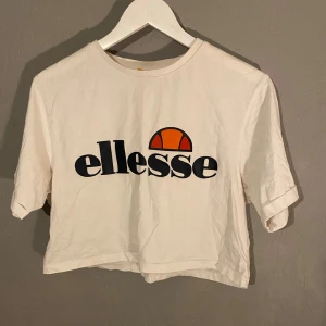 Vit croppad t-shirt från Ellesse - Säljer en snygg vit croppad t-shirt från Ellesse med deras ikoniska logga i svart och orange på bröstet. Perfekt för en avslappnad stil och passar bra till både jeans och shorts. Perfekt för sommardagar! 🌞Storlek M men passar även S (PM OM DU VILL HA FLER BILDER)