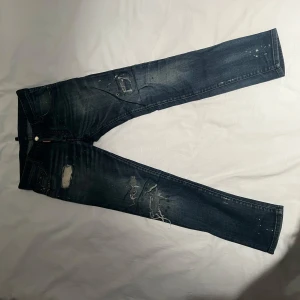 Blå jeans från Dsquared2 - Säljer ett par snygga blå jeans från Dsquared2 med coola slitningar och distressed detaljer. De har en slim passform och är perfekta för en avslappnad men stilren look. Jeansen har en klassisk femficksdesign och en läderpatch med logga baktill. Perfekta för både vardag och fest!