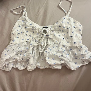 Vit blommig linnetopp - Söt vit linnetopp med små blå blommor och justerbara axelband. Toppen har en knytning framtill som ger en fin detalj och en luftig passform. Perfekt för varma sommardagar eller en avslappnad dag på stan.