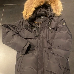 Svart dunjacka med pälsdetalj från Hollies - Säljer en svart dunjacka i storlek 12 (som en XXS) från Hollies i mycket bra skick. Endast använd ett fåtal gånger. Jackan har en mysig pälsdetalj på huvan och praktiska fickor med dragkedja. Perfekt för kalla vinterdagar! 🖤 Nypris 4500kr.