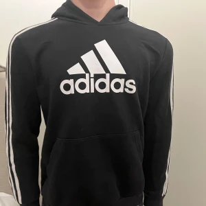 Svart hoodie från Adidas - Säljer en svart hoodie från Adidas i bra skick. Den har den klassiska loggan framtill och vita ränder längs ärmarna. Perfekt för både träning och vardag. Stor och bekväm ficka på magen. 
