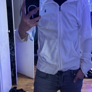 Vit hoodie från Polo Ralph Lauren - Säljer en snygg vit hoodie från Polo Ralph Lauren i storlek M. Den har en dragkedja framtill och är perfekt för både vardag och chill. Hoodien är i bra skick och har en klassisk design med det ikoniska logotypen på bröstet. Perfekt för höst och vår! Den är lite stor för mig är 190cm 72kg