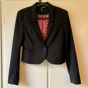 Svart kavaj från H&M - Snygg svart kavaj från H&M i bra skick. Den har en klassisk design med en knapp framtill och långa ärmar. Perfekt för både vardag och fest. Insidan har ett coolt mönster i rosa och svart.