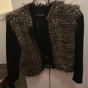 Svart jacka med fuskpäls från Rodebjer - Säljer en snygg svart jacka från Rodebjer med fuskpälsdetaljer i grått och svart. Jackan har långa ärmar och en stilren design som passar perfekt för höst och vinter. Materialet är en blandning av polyakryl och polyester, vilket gör den både varm och bekväm.