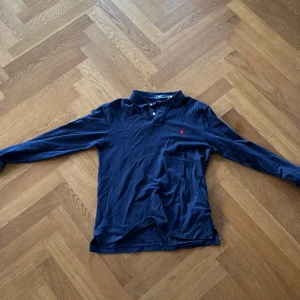 Mörkblå pikétröja från Ralph Lauren - Säljer en snygg mörkblå pikétröja från Ralph Lauren i slim fit. Tröjan har långa ärmar och en klassisk krage med knappar. Perfekt för både vardag och lite finare tillfällen. Den är i bra skick och redo för en ny ägare!