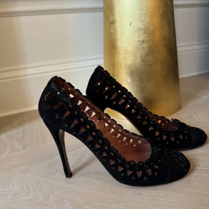 Svarta klackskor från Alaïa - Säljer ett par eleganta svarta klackskor från Alaïa i mycket bra skick. Skorna har ett unikt utskuret mönster och är tillverkade i skinn. Perfekta för festliga tillfällen med en stilren design och bekväm passform.