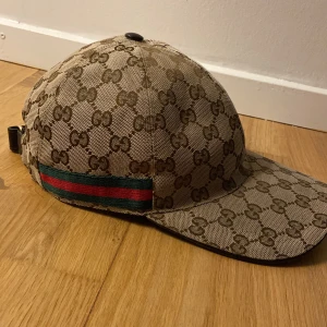 Beige keps från Gucci - Säljer en snygg beige keps från Gucci. Kepsen har en justerbar rem baktill och en grön och röd rand på sidan. storlek M. Bra skick! Skriv för frågor.