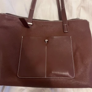 Brun axelväska från Rosetti - Säljer en snygg brun axelväska från Rosetti i skinn. Väskan har en praktisk dragkedja upptill och en framficka med knappdetalj. Perfekt för vardagsbruk och rymmer allt du behöver. Den är i mycket bra skick och redo för en ny ägare!