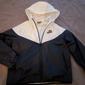 Svart och vit vindjacka från Nike - Säljer en snygg svart och vit vindjacka från Nike. Jackan har en dragkedja framtill och en huva för extra skydd mot väder. Perfekt för höst och vår! Den är i mycket bra skick och passar perfekt för både träning och vardag.