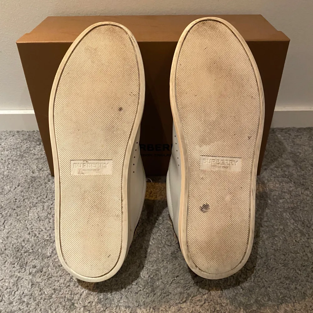 Säljer ett par snygga vita sneakers från Burberry i mycket bra skick. De har en klassisk design med beiga detalger och den ikoniska Burberry-rutiga baksidan. Perfekta för både vardag och fest! Storlek är 42,5 men passar även 42.. Skor.