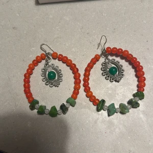 Färgglada örhängen med pärlor och stenar - Säljer ett par unika och färgglada örhängen med röda pärlor och gröna stenar. De har en cool design med en silverfärgad detalj i mitten som ger en bohemisk känsla. Perfekta för att liva upp vilken outfit som helst! 🌟