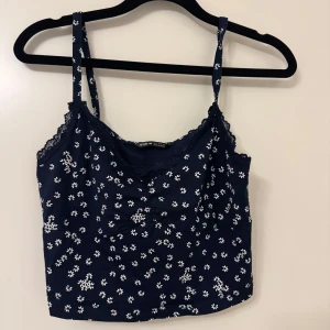 Marinblå blommig linnetopp - Söt marinblå linnetopp med vita blommor. Toppen har smala axelband och spetsdetaljer vid halsringningen. Använd en enstaka gång säljer då den blivit för liten.