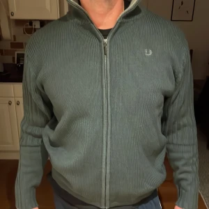 Fred Perry stickad tröja - Säljer en snygg grön ribbad tröja med dragkedja framtill. Tröjan är i storlek XXL men skulle säga att den passar mindre. Perfekt för höst och vår, med långa ärmar och en bekväm passform. Den har en diskret logga på bröstet. Passar både till vardags och lite mer uppklädda tillfällen. Modellen är 186 och väger ~90kg, lite tajt på honom dock.