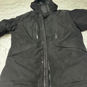 Svart dunjacka från Sail Racing - Säljer en svart dunjacka från Sail Racing, perfekt för kalla vinterdagar. Jackan har en cool logga på ärmen och flera praktiska fickor med dragkedja. Den är i mycket bra skick och har en skön, vadderad insida för extra värme. Passar perfekt för vinteräventyr eller bara för att hålla sig varm i stan.