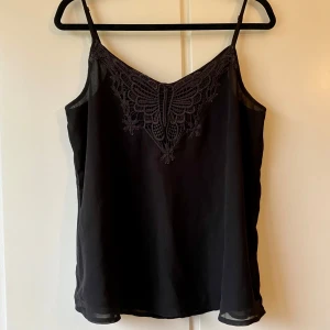 Svart linne med spetsdetaljer - Snyggt svart linne med tunna axelband och vacker spetsdetalj framtill. Perfekt för både vardag och fest. Materialet känns lätt och luftigt. 