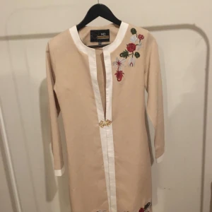 Beige kappamed broderade blommor - Säljer en elegant beige klänning från ND med vackra broderade blommor i rött och grönt. Kappan har långa ärmar och en stilren design med en dekorativ detalj framtill. Perfekt för vår och sommar!