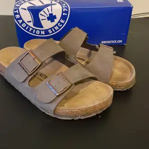 Säljer ett par klassiska Birkenstock sandaler i brunt skinn. De har två justerbara remmar med metallspännen och en bekväm korksula. Perfekta för sommaren och ger bra stöd för fötterna. Använda men i bra skick. Modell arizona.