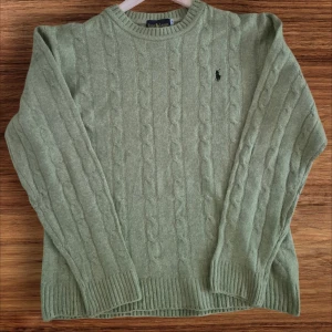 Grön stickad tröja från Ralph Lauren - Säljer en snygg grön stickad tröja från Ralph Lauren. Den har ett klassiskt kabelstickat mönster och en liten logga på bröstet. Perfekt för kyliga dagar och passar både till vardags och lite finare tillfällen. Tröjan har långa ärmar och ribbade muddar för extra komfort.