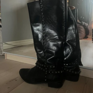 Svarta skinnstövlar med nitar - Säljer ett par svarta skinnstövlar i mycket bra skick. De har coola nitar längs skaftet och runt fotleden, vilket ger dem en edgy look. Perfekta för höst och vinter, och de passar bra till både vardag och fest. 