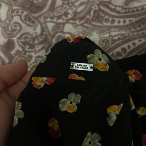 Blommig klänning från Armani Exchange - Säljer en superfin svart klänning från Armani Exchange med färgglada blommor i rött, gult och grönt. Klänningen har långa ärmar med volangdetaljer och en bekväm passform. Perfekt för både vardag och fest!