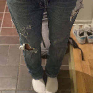 Blå jeans med slitningar - Säljer ett par blå jeans med coola slitningar på knäna. De har en normal passform och är perfekta för en avslappnad stil. Jeansen är i bra skick och passar perfekt till vardagsoutfiten.