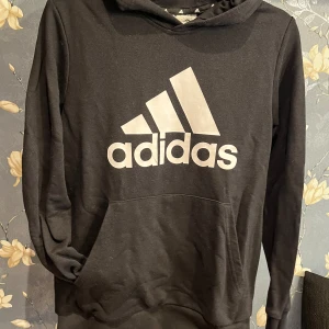 Svart hoodie från Adidas - Säljer en svart hoodie från Adidas i storlek S Den har en stor vit logga på framsidan och är perfekt för både träning och vardag. Materialet är mjukt och bekvämt, och den har en klassisk passform med långa ärmar och huva. Perfekt för höst och vinter!