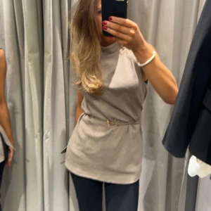 Beige ärmlös topp med bälte - Snygg beige ärmlös topp med hög krage och ett elegant bälte i midjan. 