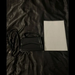 Svart mini handväska  - En mindre handväska som används få tal gånger! (Mäter den genom ett A4 papper på bild så man ser ungefär hur stor den är😊) axelband medföljer!