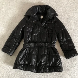 Hugo Boss svart glansig pufferjacka med skärp - Otroligt snygg lätt glansig pufferjacka från Hugo Boss med trekvartsärmar och skärp i midjan. Väldigt varm och passar utmärkt för höst och vintersäsongen. Endast använd ett fåtal gånger och är i nyskick, utan anmärkningar. Ordinariepris cirka 3500SEK