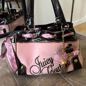 Juicy couture väska - Säljer min Vintage rosa/ brun Juicy couture väska med mycket detaljer och tillbehör. Den har några år på naken därav är den ganska sliten men mest på kanterna som syns på bilderna. Handtaget ser fint ut fortfarande. Väskan har två små fack på sidorna