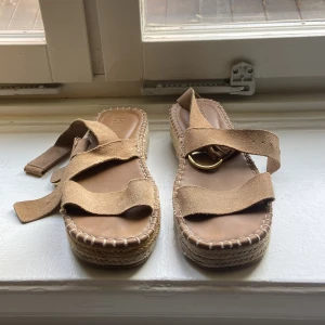 Beige sandaler från Zara - Säljer ett par snygga och bekväma sandaler från Zara i storlek 38. De är i en fin beige färg med en stilren design. Sandalerna har en platåsula med flätad detalj och justerbara remmar i mocka med guldfärgade spännen. Perfekta för sommaren!