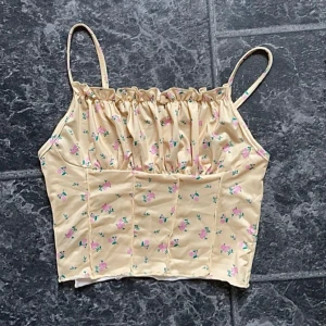 Beige blommig korsettopp - Säljer en superfin créme gul topp med små blommor i rosa och grönt. Toppen har smala axelband och en söt rynkad detalj vid bysten. Perfekt för sommardagar eller en festlig kväll!