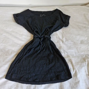Svart klänning från Forplay - Säljer en snygg svart klänning från Forplay i storlek S. Den är tillverkad av 100% bomull vilket gör den superbekväm och perfekt för vardagsbruk. Klänningen har korta ärmar och ett knytband i midjan för en fin passform. En av öglorna som bandet träs igenom är sliten, annars är den i fint skick. Perfekt för både avslappnade dagar och festligare tillfällen!