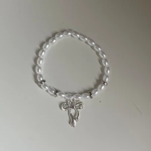 Pärlarmband med silverfärgad rosett - Säljer ett superfint armband med vita pärlor och en söt silverfärgad rosett som hänge. Perfekt för att ge en elegant touch till vilken outfit som helst. Armbandet är elastiskt och passar de flesta handleder. Perfekt för både vardag och fest! ✨