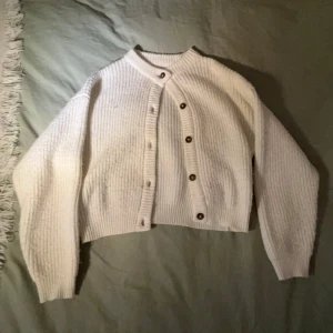 Vit stickad kofta med knappar - Säljer en supermysig vit stickad kofta med bruna knappar. Den är croppad och har långa ärmar, perfekt för höst och vinter. Koftan är i nyskick och passar både till vardags och lite finare tillfällen. Perfekt att matcha med jeans eller en kjol!