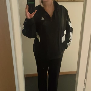 Svart vindjacka från Adidas - Säljer en svart vindjacka från Adidas med vita detaljer och de klassiska tre ränderna på ärmarna. Jackan har en dragkedja framtill, en ficka på höger sida och är perfekt för både träning och vardag. Den är i nyskick. Köpt i Danmark och är en limited edition.