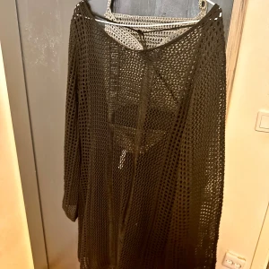 Svart stickad klänning - Säljer en superfin svart stickad klänning med ett genombrutet mönster. Den har en tight passform och är perfekt för festliga tillfällen. Klänningen är långärmad och har en djup v-ringning i ryggen med snörning. Perfekt för att styla med ett par snygga klackar!