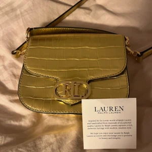 Guldig axelväska från Ralph Lauren - Säljer en superfin guldig axelväska från Ralph Lauren. Väskan är i skinn med ett snyggt krokodilpräglat mönster och har en elegant RL-logga på framsidan. Perfekt för att ge lite lyxig touch till din outfit! Väskan har aldrig använts utan bara varit upp i garderoben 
