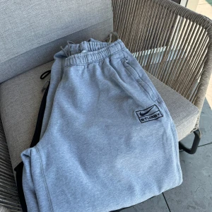 Grå mjukisbyxor från Nike x Stussy - Säljer ett par grå mjukisbyxor från Nike i samarbete med Stussy. De är i storlek L och har en broderad logga på sidan. Byxorna har en elastisk midja med snörning och är superbekväma för chill eller träning. Perfekta för höst och vinter!