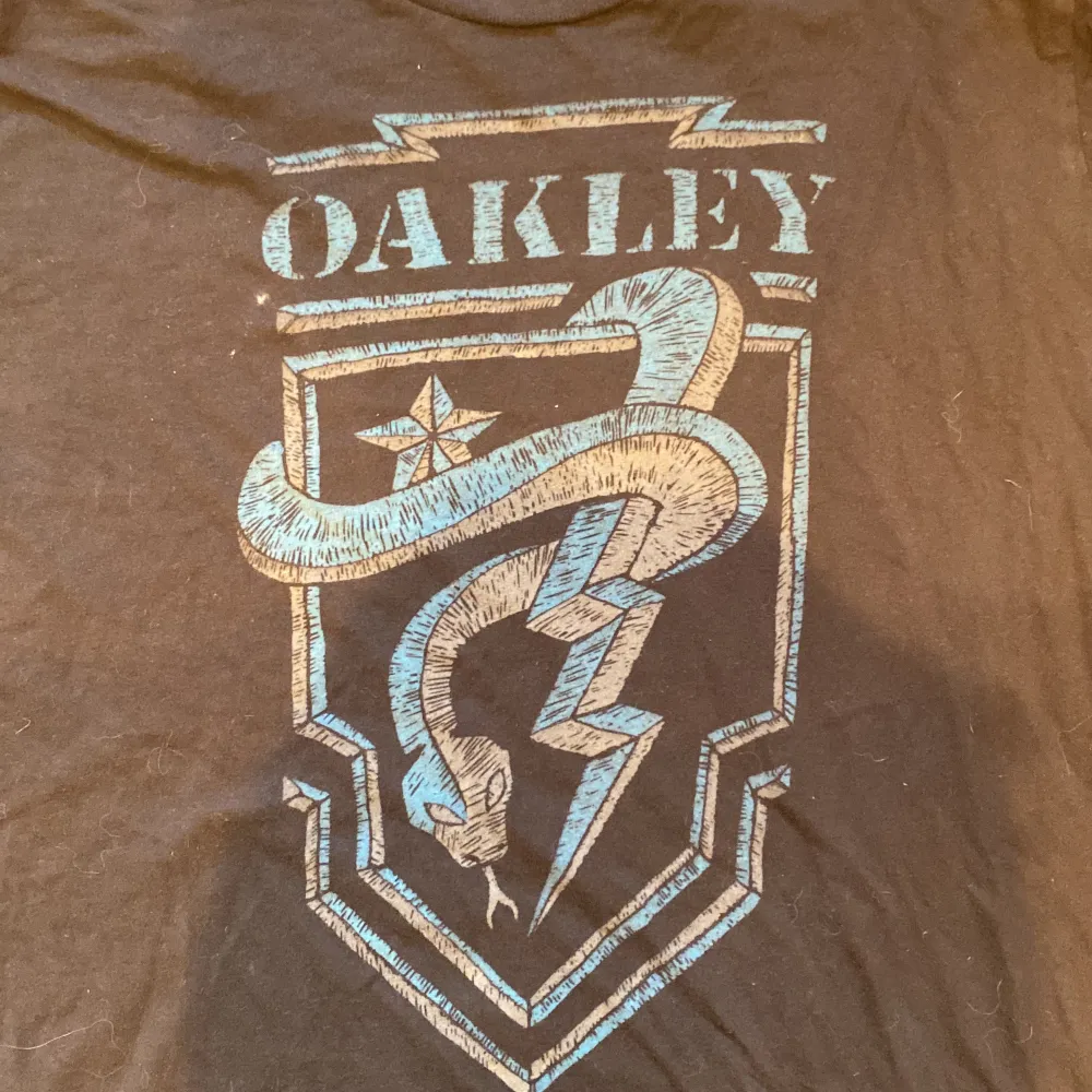 Säljer en svart t-shirt från Oakley med ett coolt tryck i blått och vitt på framsidan. T-shirten har en normal passform och är tillverkad i bomull, vilket gör den bekväm att bära. Perfekt för vardagsbruk eller träning! Storlek XL men sitter som M. T-shirts.