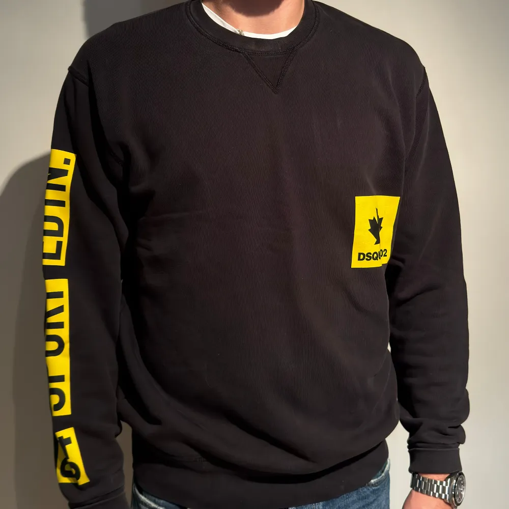 Säljer en svart sweatshirt från Dsquared2 med coola gula detaljer. Tröjan har en gul logga på bröstet och text längs ärmen. Perfekt för en sportig look eller bara för att chilla i. Den är långärmad och gjord i ett bekvämt material. Passar perfekt till jeans eller joggers!. Tröjor & Koftor.