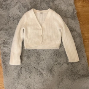 Vit stickad kofta från Lindex - Säljer en superfin vit stickad kofta från Lindex i storlek 146/152. Den har en v-ringad design med knappar framtill och är croppad. Perfekt för höst och vinter, och den är både mysig och stilren. 